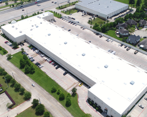 Aerial view of a large white industrial warehouse with surrounding parking lots, nearby roads, and adjacent smaller buildings.