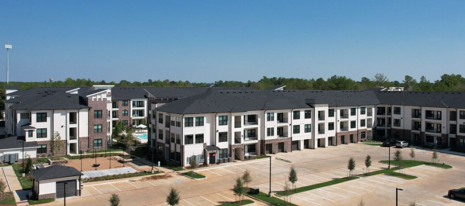 Wide view of a modern three-story apartment complex with parking lot and landscaped greenery under a clear blue sky.
