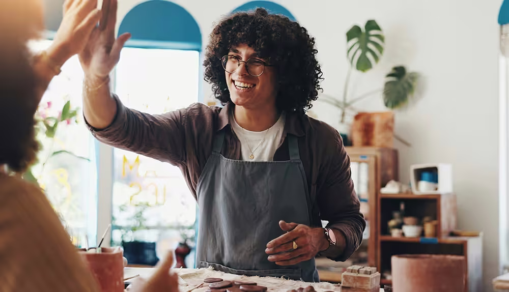 A person with curly hair wearing an apron giving a high five in a bright craft studio