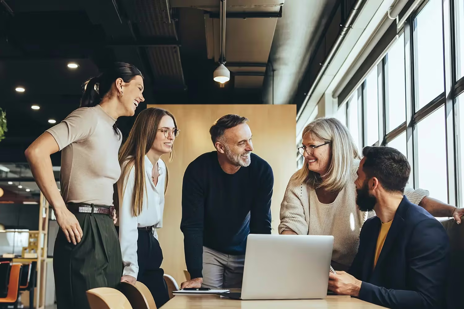 A diverse group of five colleagues laughing together around a laptop in a modern office
