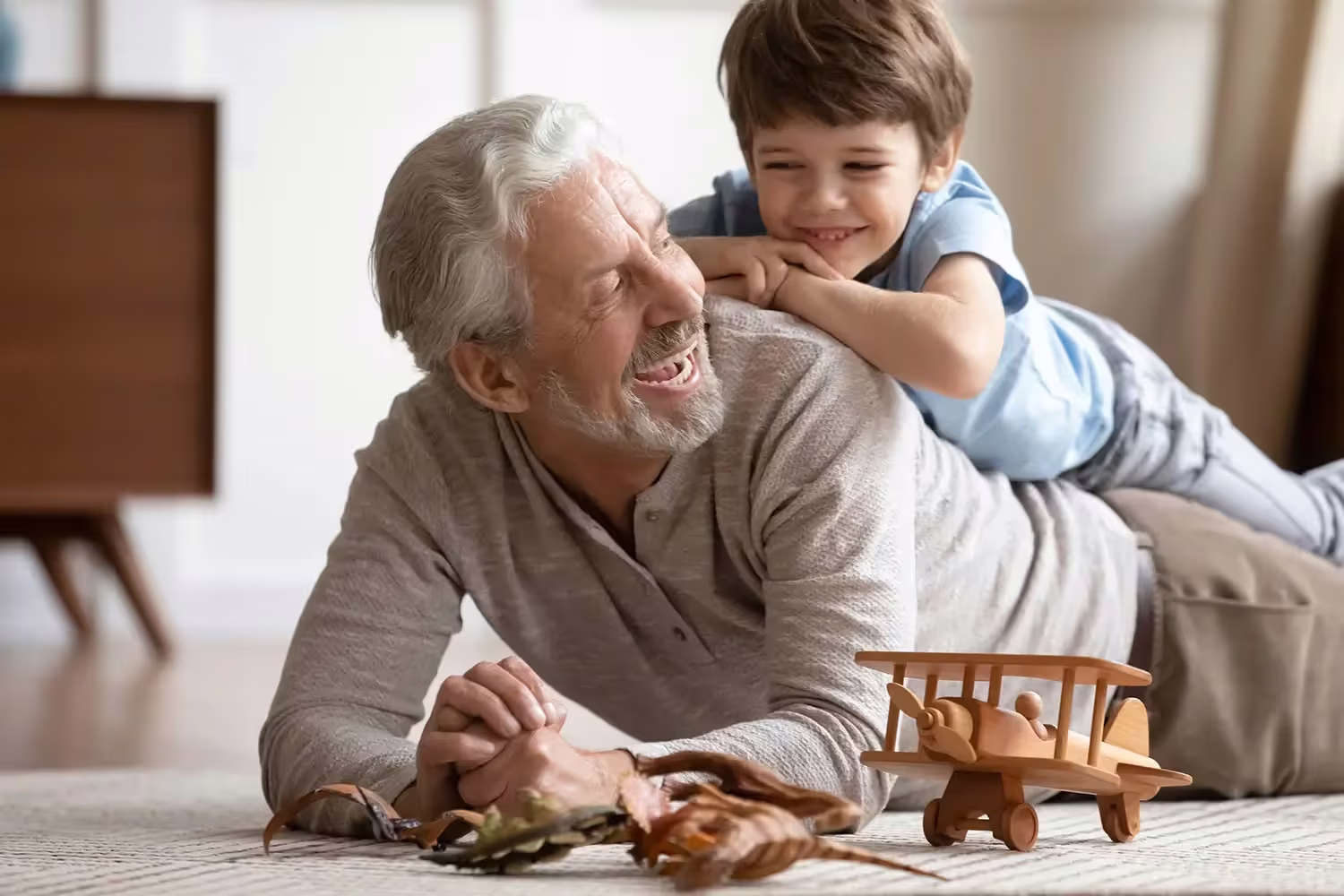 A grandfather lying on the floor playing with his young grandson with a wooden toy aeroplane