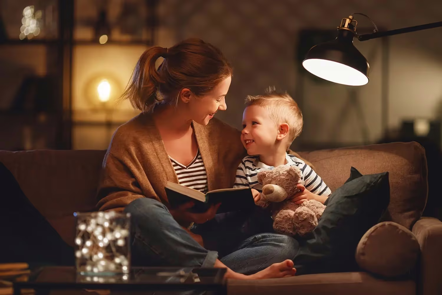 A mother reading a book to her young son on a sofa by warm lamplight, both smiling