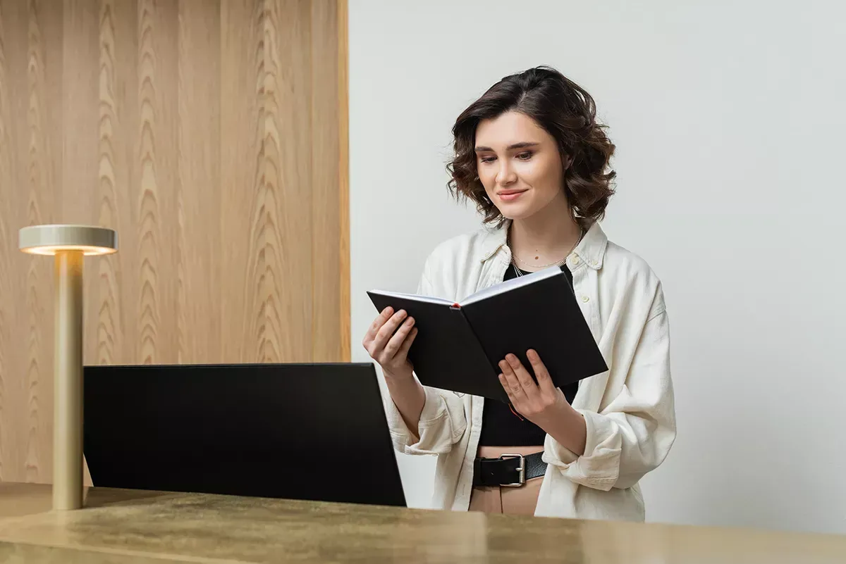 Young woman reading a black notebook while standing at a desk with a laptop and a lamp.