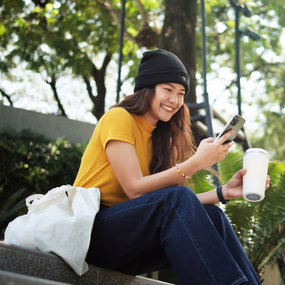 Young woman in a black beanie and yellow shirt sitting outside, smiling at her phone and holding a white tumbler.