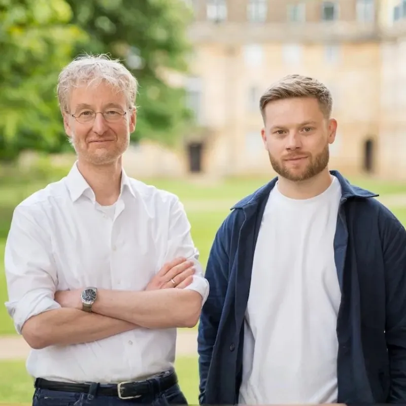 Two men standing outside with green trees and a blurred building in the background, one wearing glasses and a white shirt with arms crossed, the other in a white t-shirt and dark jacket.