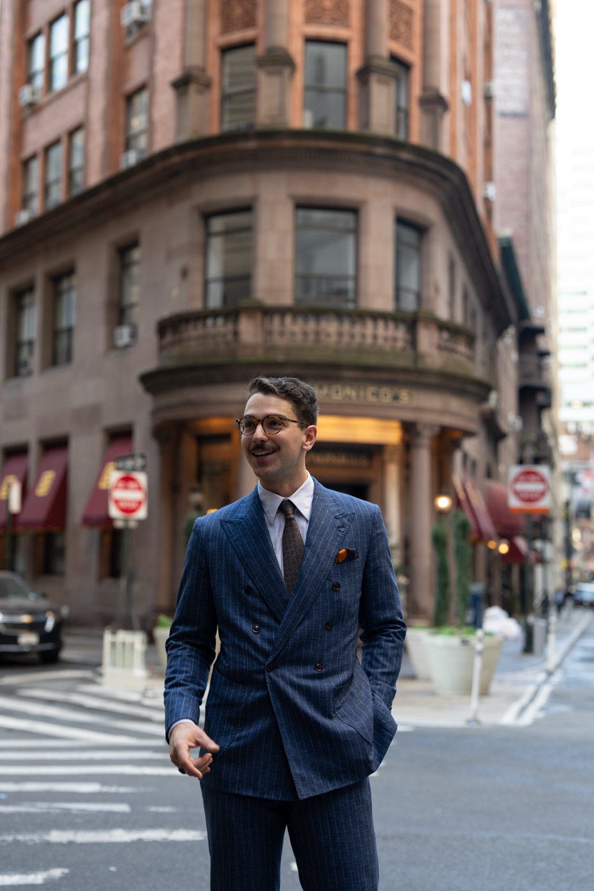 Man in a blue pinstripe suit with glasses standing on a city street crosswalk in front of a historic building.