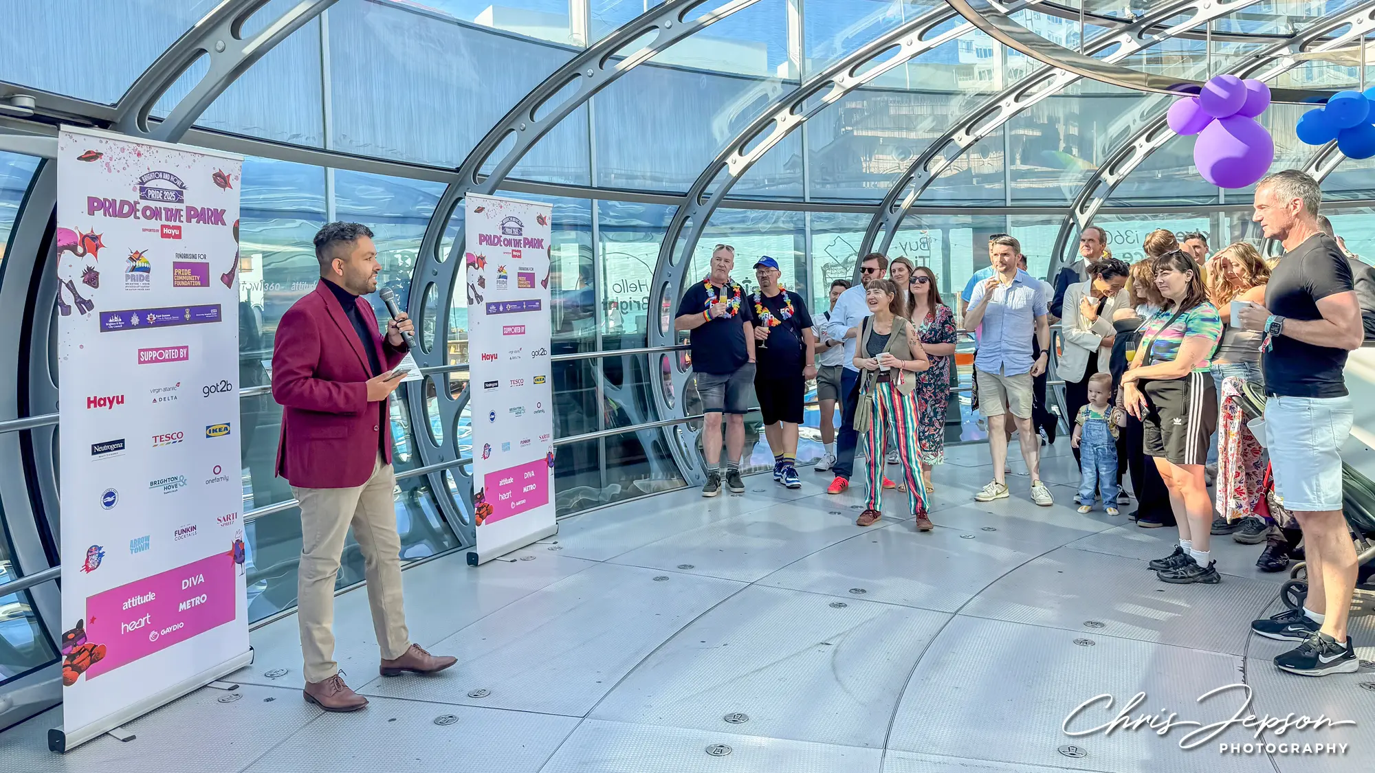 Leslie in a burgundy blazer speaking into a microphone to a diverse audience in a glass-enclosed space with Pride on the Park banners.