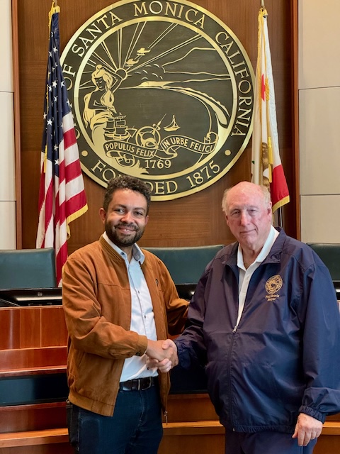 Leslie Clarke and Phil Brock, Mayor of Santa Monica, shaking hands in front of a large City of Santa Monica seal with American and California state flags behind them.