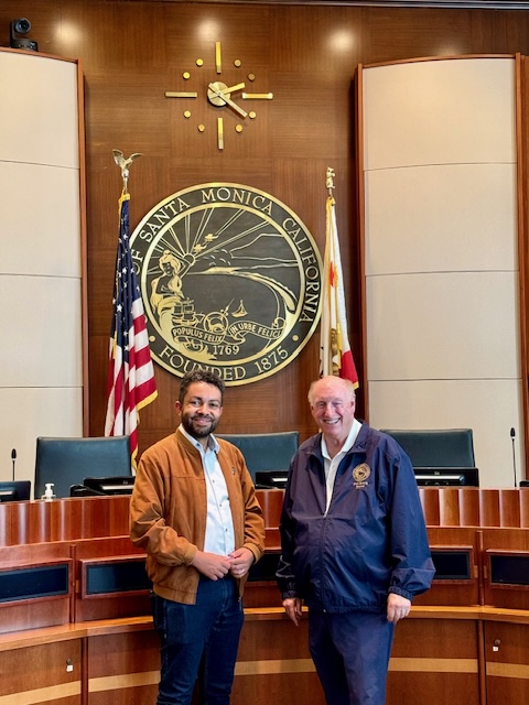 Leslie and Phil Brock, Mayor of Santa Monica, standing and smiling in front of a Santa Monica, California city seal with American and Californian flags in a wood-paneled official meeting room.