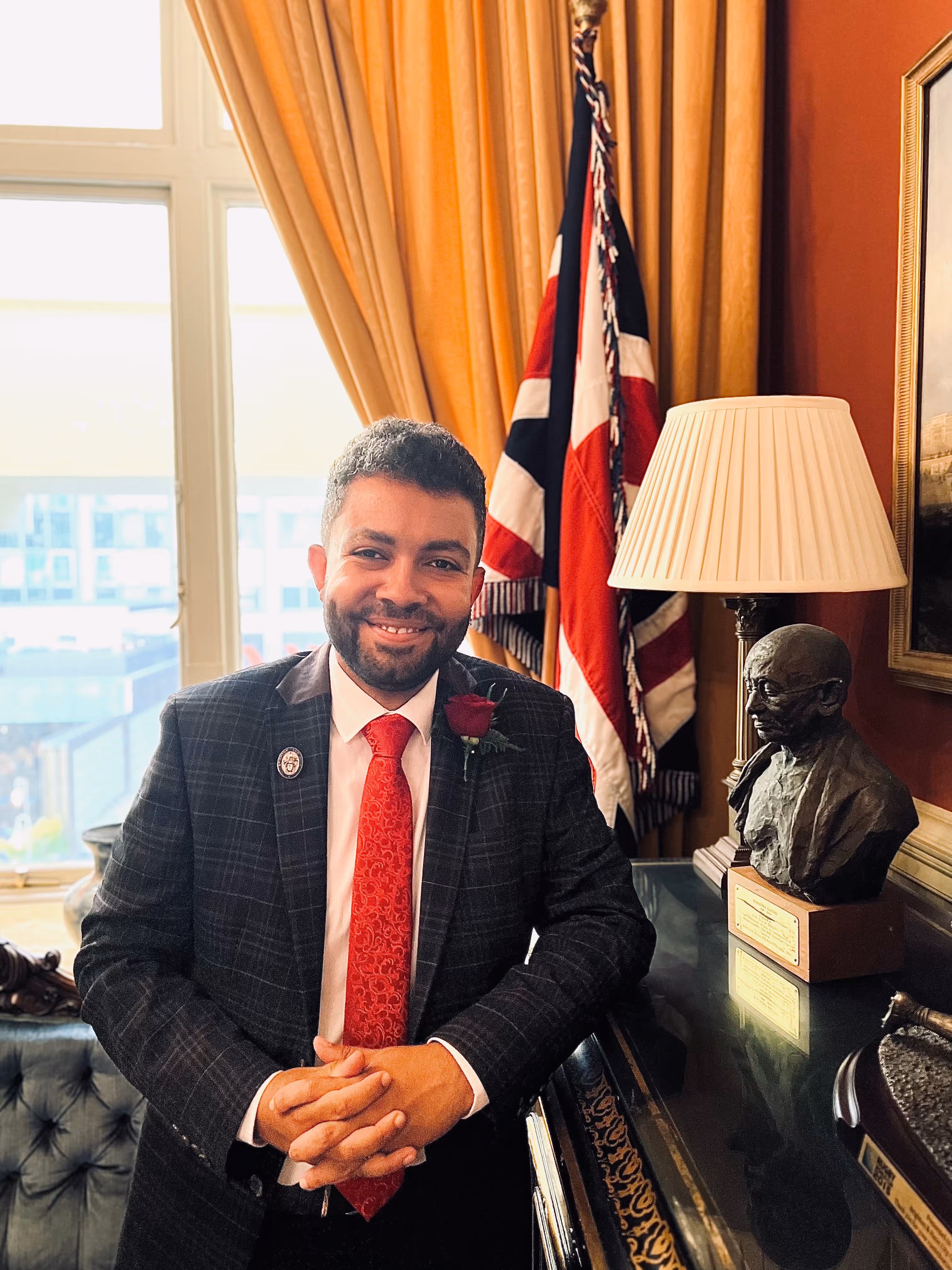 Leslie, smiling in a dark plaid suit and red tie standing in a room with beige curtains, a British flag, a bust statue, and a lamp.