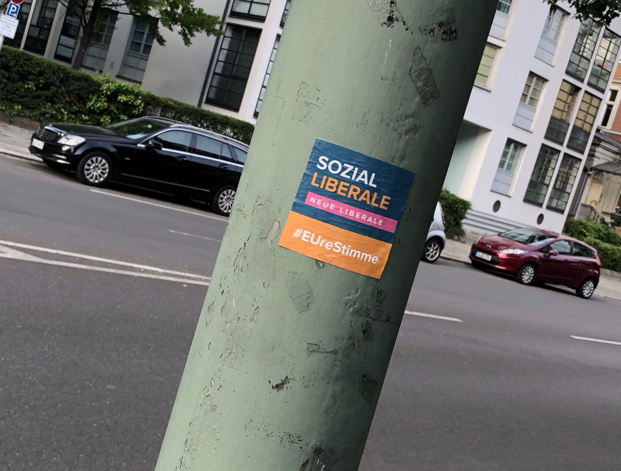 Sticker on a green pole with the text 'SOZIAL LIBERALE NEUE LIBERALE #EUreStimme' on a city street with parked cars and buildings in the background.