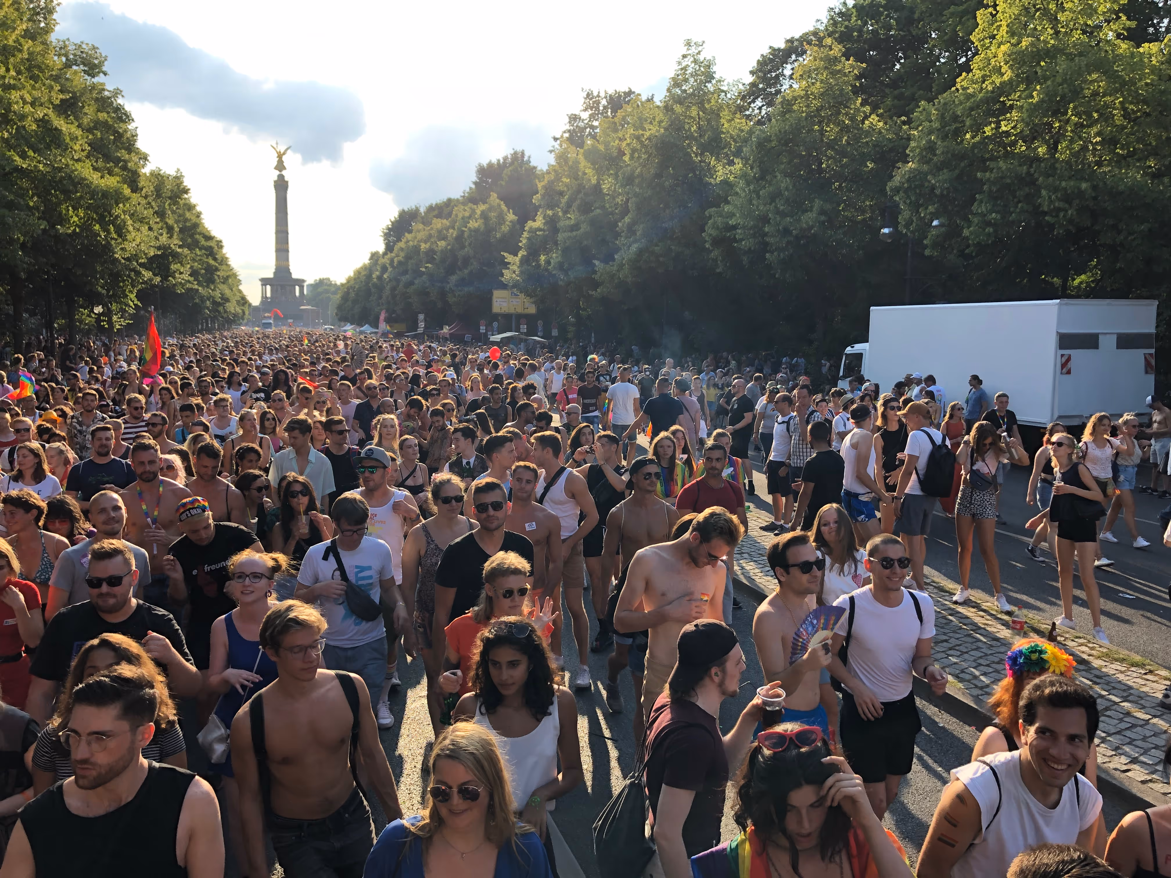 Large crowd of people walking behind dopestudi0s' Pride float on a sunny day near the Victory Column monument in Berlin.