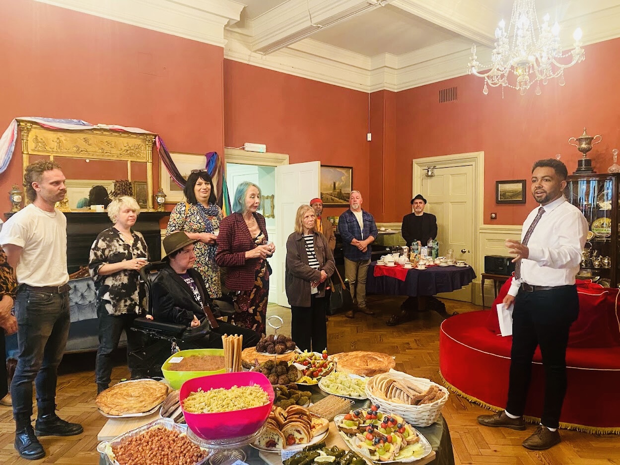 A group of people from the TNBI community, councillors and council officers standing and listening to Leslie speaking in the Mayor's Parlour, a room with red walls, a chandelier, and tables with various foods and drinks.