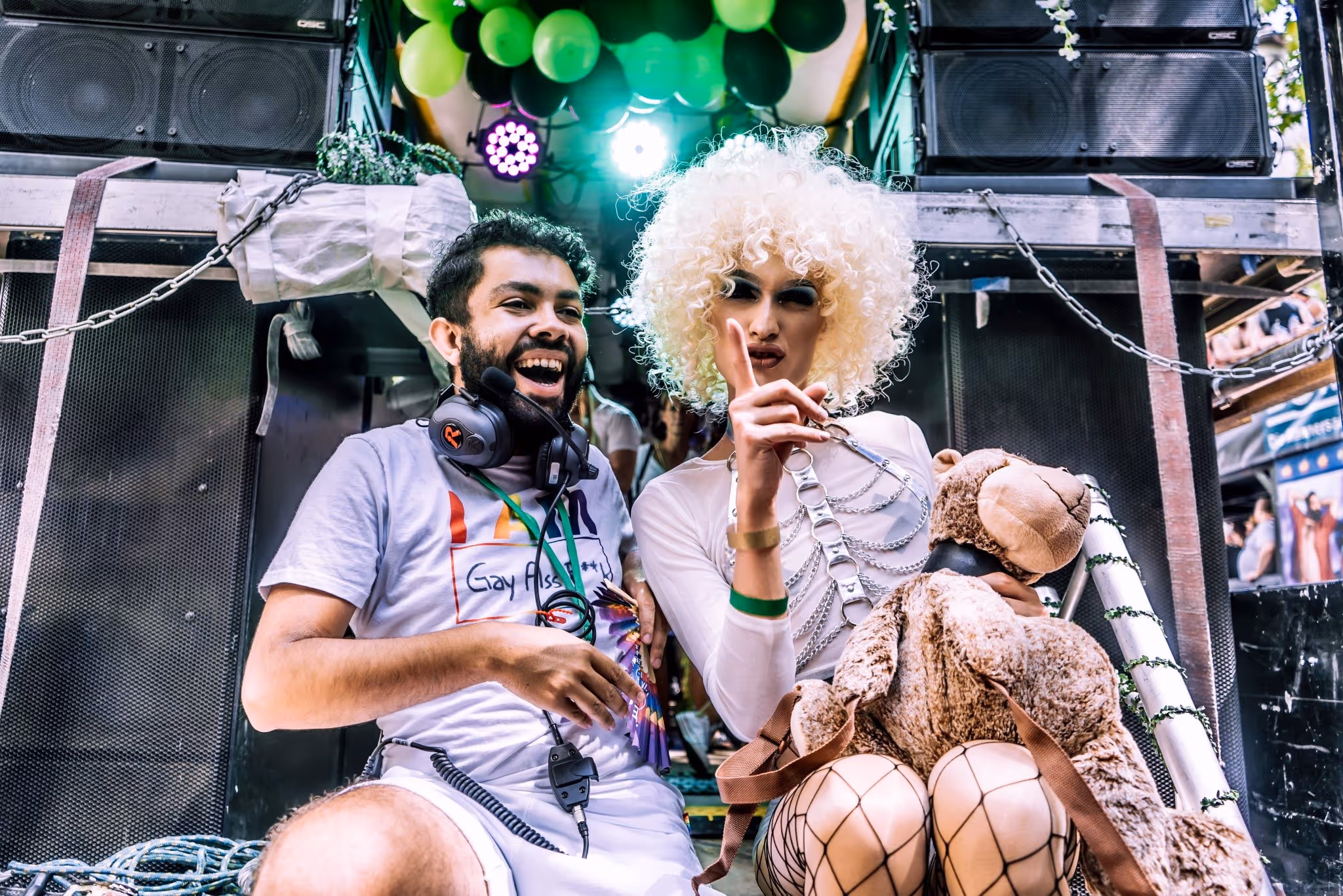 Leslie and Queen Amy Strong sitting and smiling; one wearing a headset and a shirt with rainbow text, the other with curly blonde hair, fishnet stockings, chains, and holding a teddy bear.