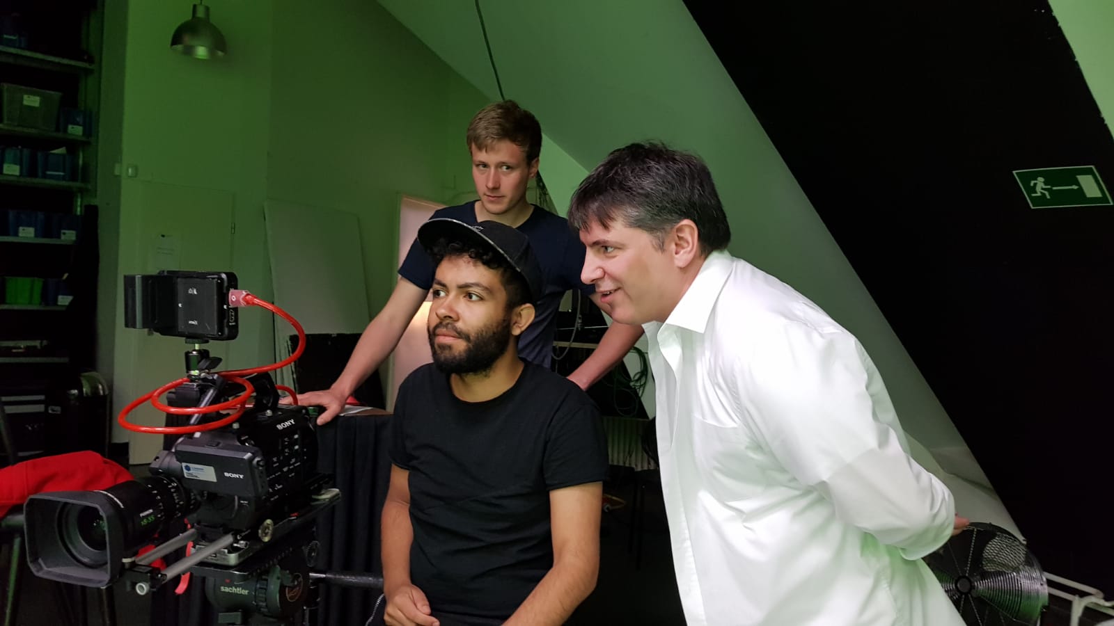 Leslie, a camera operator and the Spitzenkandidat for the Social Liberals watching a camera monitor on a film set, one sitting and two standing behind.