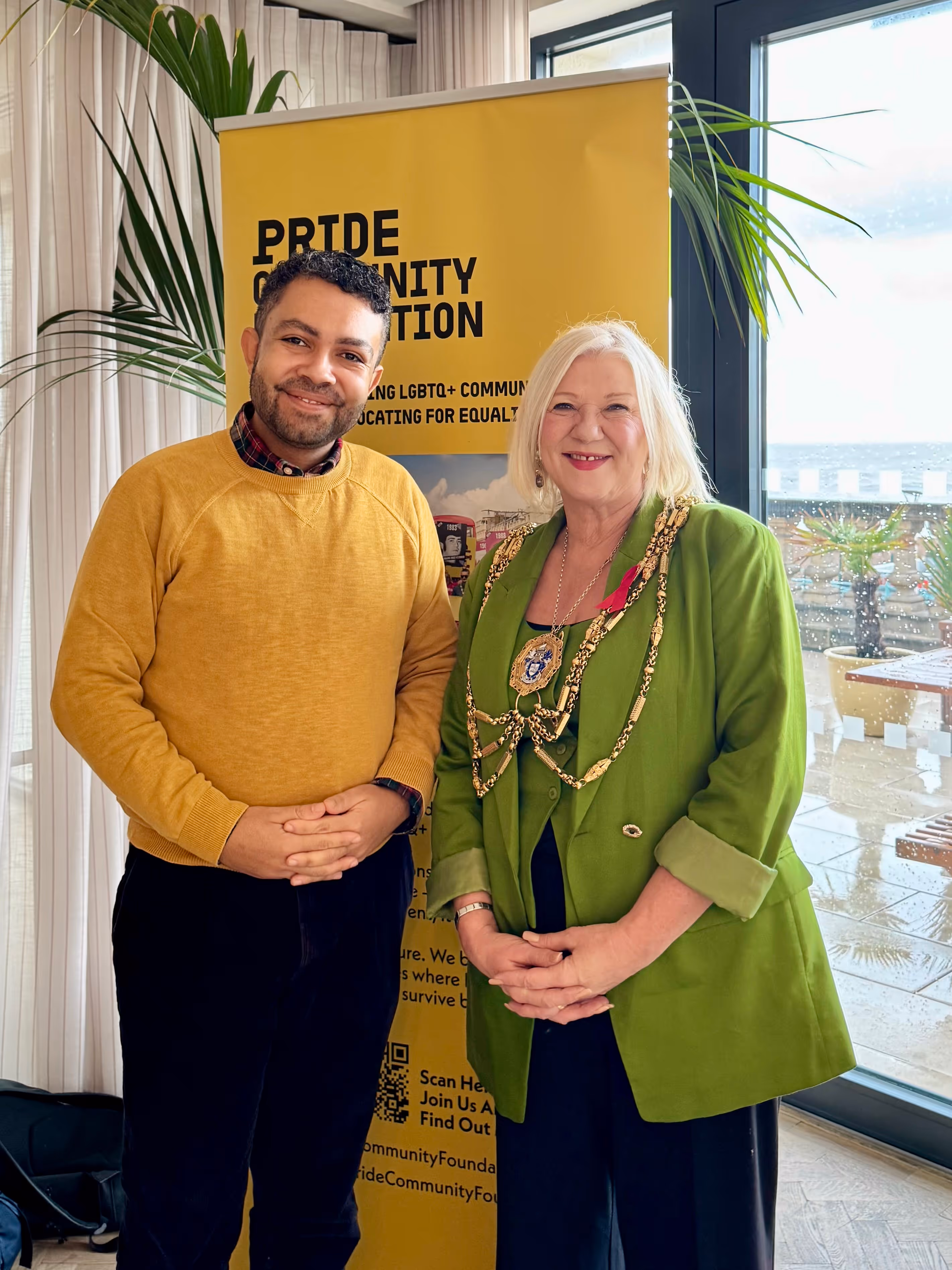 Leslie in a mustard yellow sweater and Cllr Amanda Grimshaw, Mayor of Brighton & Hove, wearing a green blazer with a ceremonial chain stand smiling indoors in front of a yellow Pride Community Foundation banner.