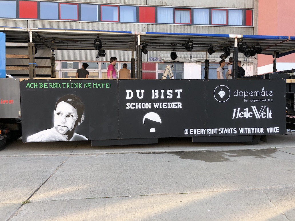 Outdoor black banner on truck with a stencil portrait of a AfD politicians' face, green text saying 'ACH BERNDT TRINK NE MATE!', white text 'DU BIST SCHON WIEDER', logos for dopemate and Heile Welt, and the phrase 'EVERY RIOT STARTS WITH YOUR VOICE.'