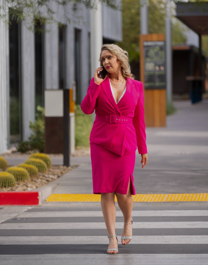 Woman in a bright pink belted blazer dress walking on a crosswalk while talking on a smartphone.
