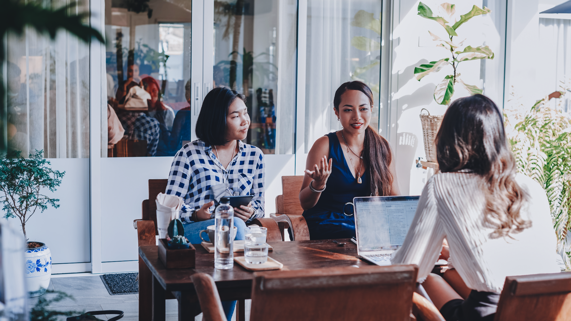 Smiling woman with curly blonde hair wearing a light blazer, typing on a laptop at a desk with flowers and framed photos in the background.