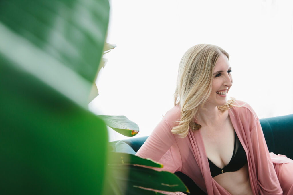 Blond woman in black bra and pink robe sitting on a teal sofa.  Single women should consider boudoir photography as an act of self-love.  Photography by Lindsay Hite