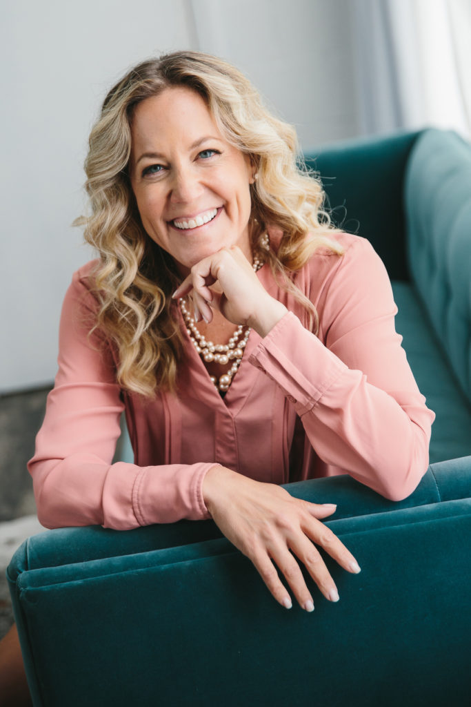 Headshot of blonde woman in pink blouse on teal sofa, by Lindsay Hite