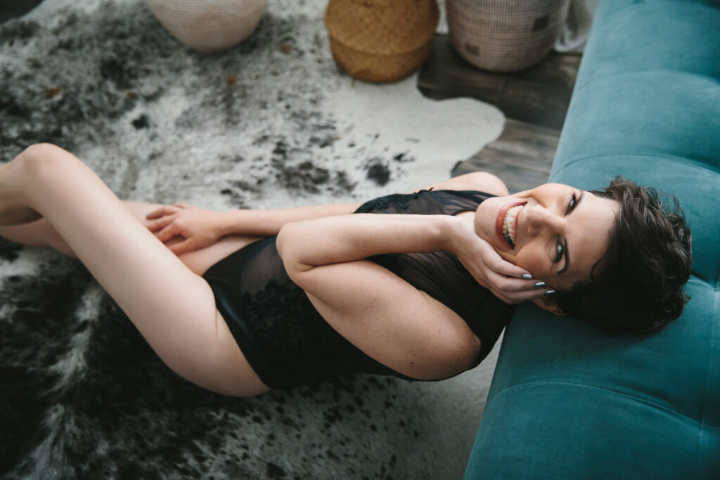 Woman with short hair in black lingerie, sitting on the floor and leaning back again a teal sofa.  Photography by Lindsay Hite