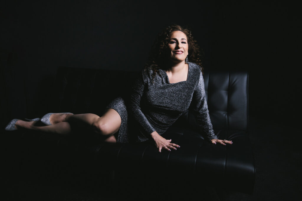Woman in a black sparkly dress with silver heals sitting on a black sofa with a black background. Boudoir photography by Lindsay Hite