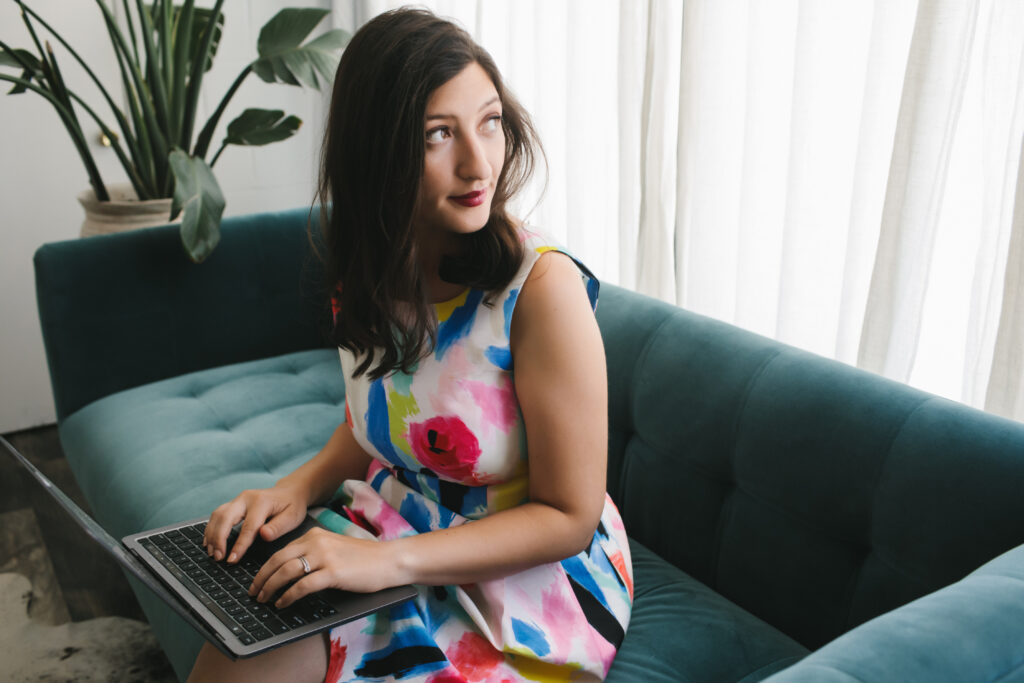 Woman in floral dress working at her laptop sitting on a teal sofa.    Branding photography by Lindsay Hite at Show Your Spark. 