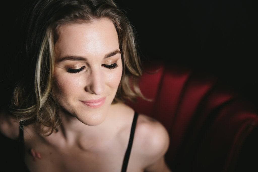Woman with a peaceful smile on her face and her eyes closed sitting on a red velvet chair with a dark background.  Boudoir photography by Lindsay Hite.