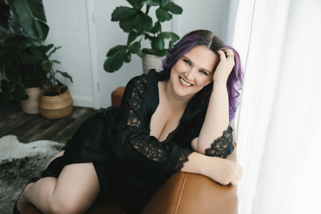 Woman wearing a lacy black dress sitting on a leather sofa with a white backdrop and plants in the backdrop. Photography by Lindsay Hite at Show Your Spark Braintree, MA.