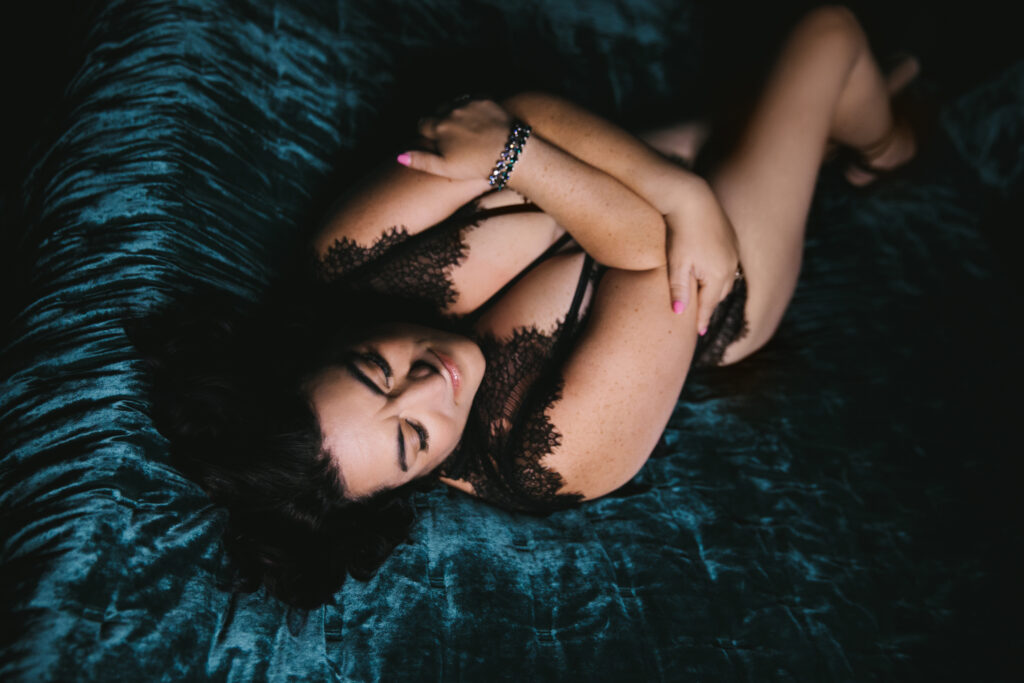 Woman in a black lace body suit in a dark room on her back on a dark teal jewel-toned bed declaring her fierceness. Photography by Lindsay Hite
