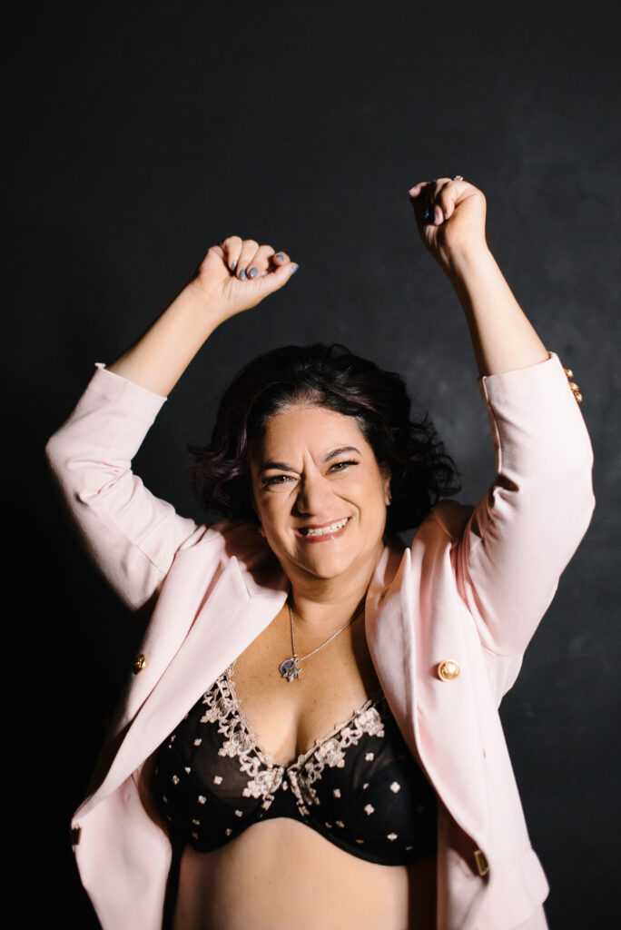 Woman in a black bra and pink jacket smiling with a grey background. Photography by Lindsay Hite