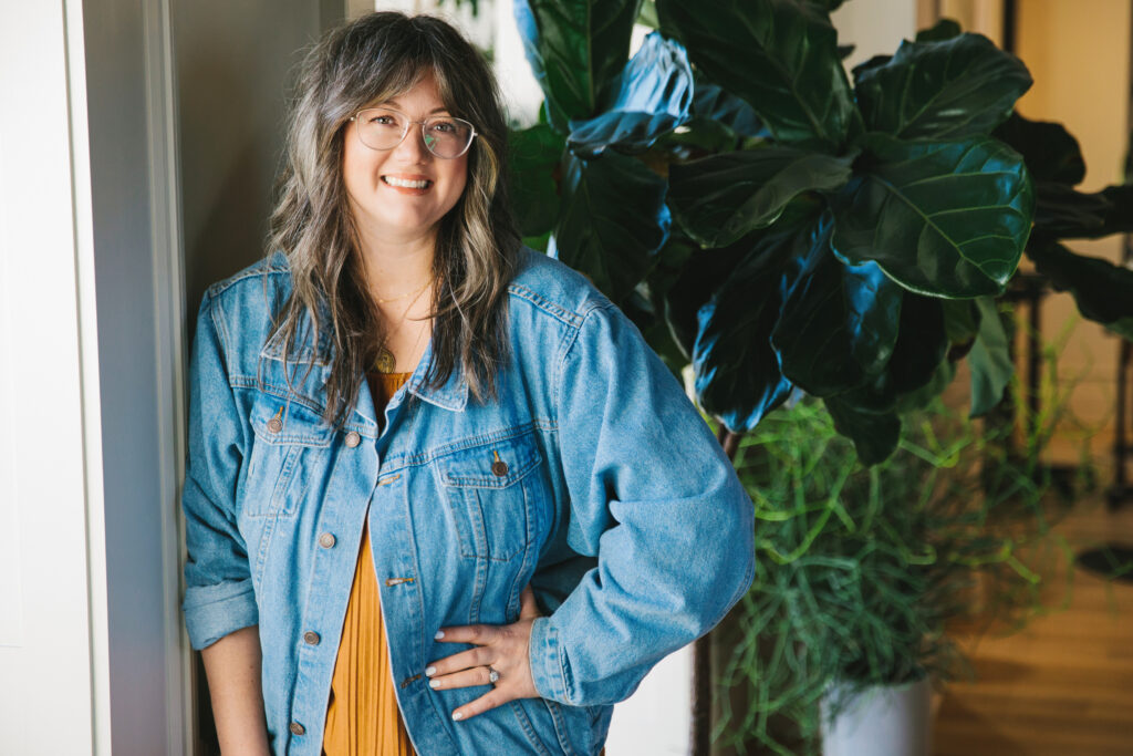 Casual headshot of a woman wearing an ochre colored dress with a denim jacket. Photography by Lindsay Hite.