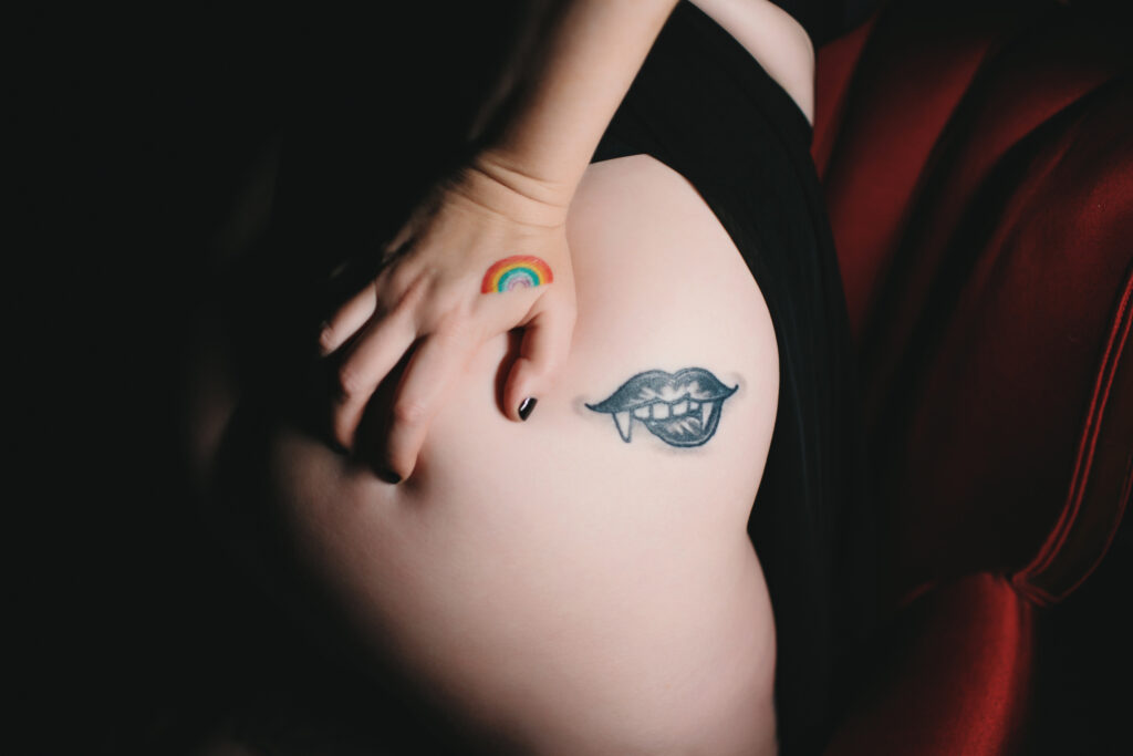 Woman leaning against a red velvet chair in black lingerie with tattoo on her hip and hand