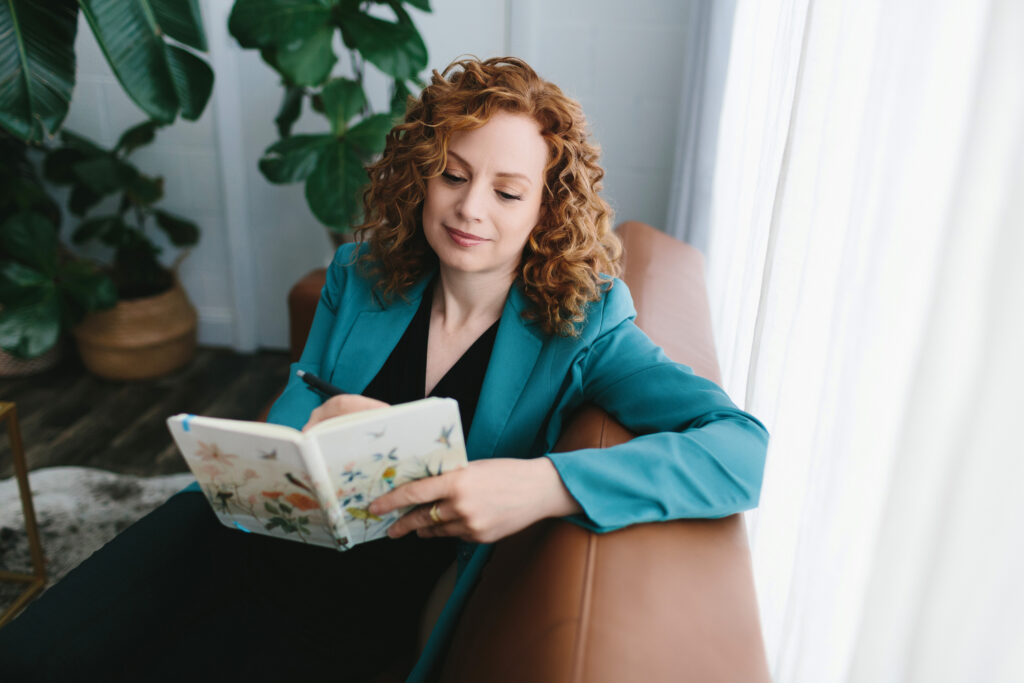 Woman in black blouse and teal jacket sitting on a leather sofa with a journal. writing about what she's learned lessons from adversity in our Fierce After Forty project by Lindsay Hite. 
