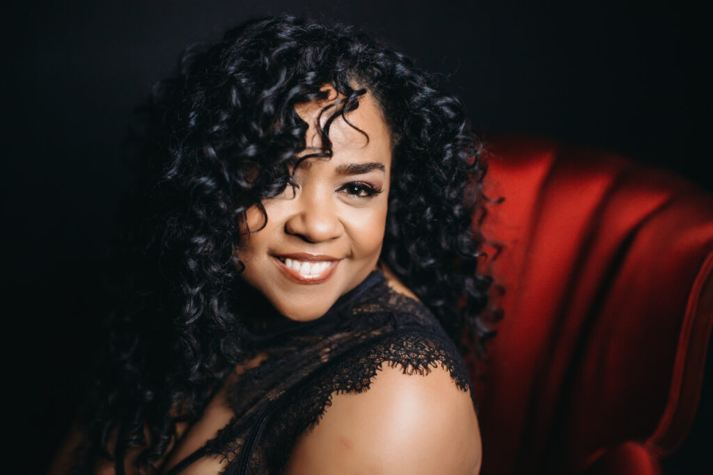 African American woman in black top sitting in a red velvet chair with a black background. Photography by Lindsay Hite