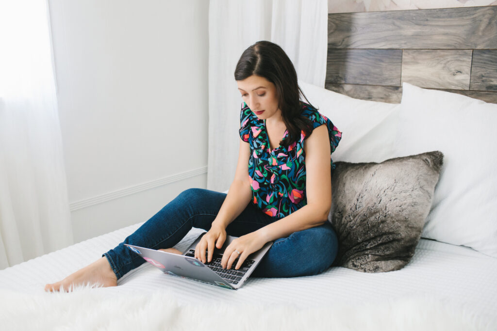 Branding session with a woman sitting on a white bed working on her laptop. Photography by Lindsay Hite.