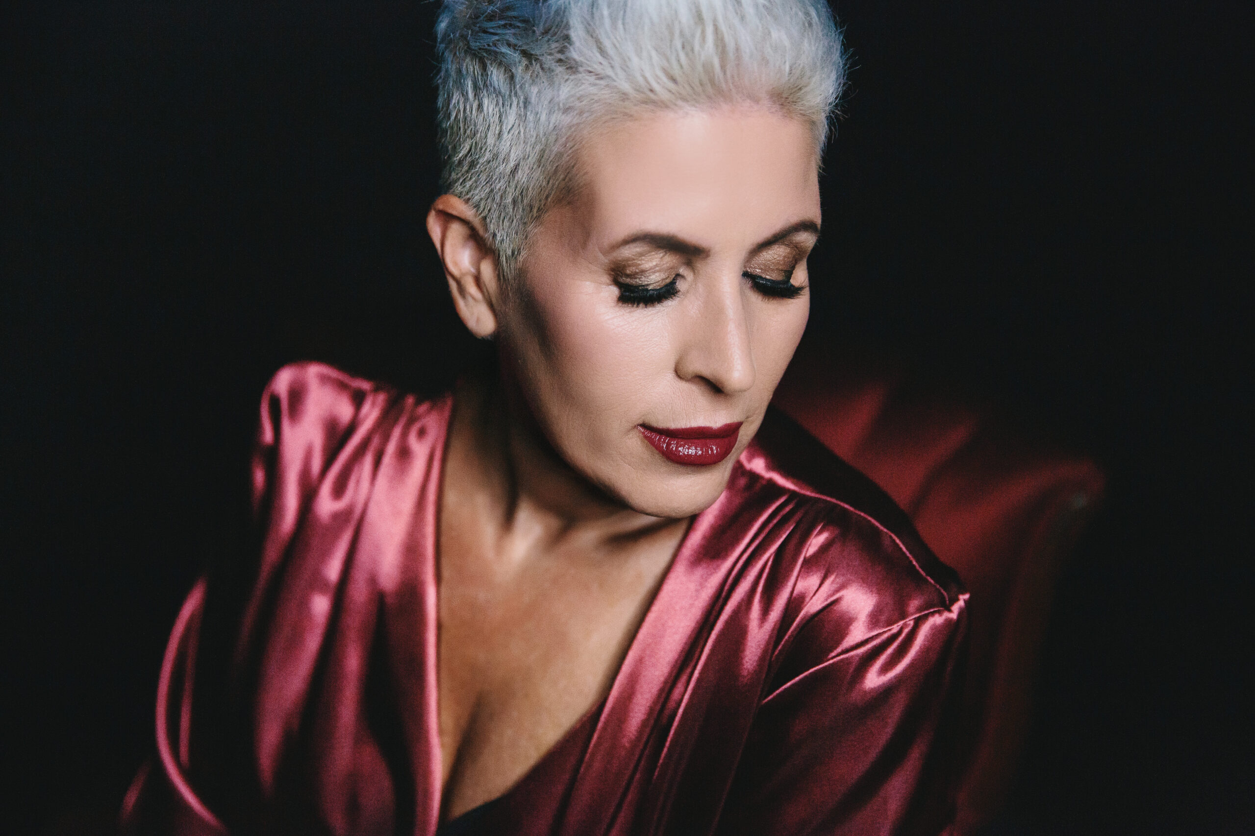 Woman in satin robe sitting in a red velvet chair with a black background.  Photography by Lindsay Hite