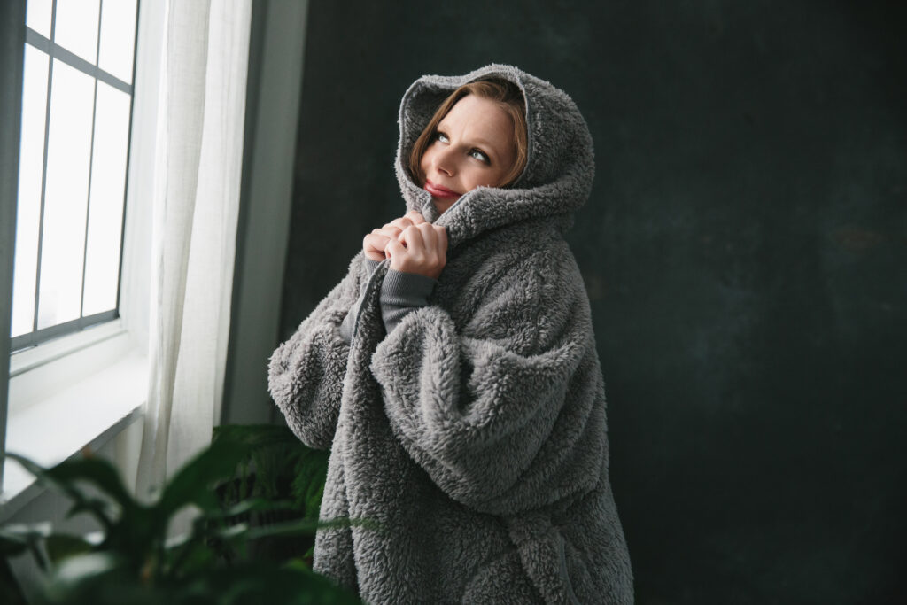 Woman in fleece hoodie blanket looking out the window with a smile on her face. Photography by Lindsay Hite