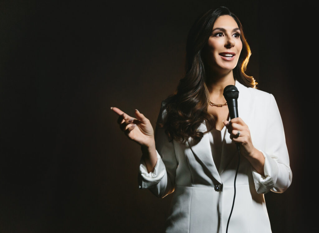 Woman in a white blazer and power suit standing in front of a dark backdrop with a microphone. Photography by Lindsay Hite