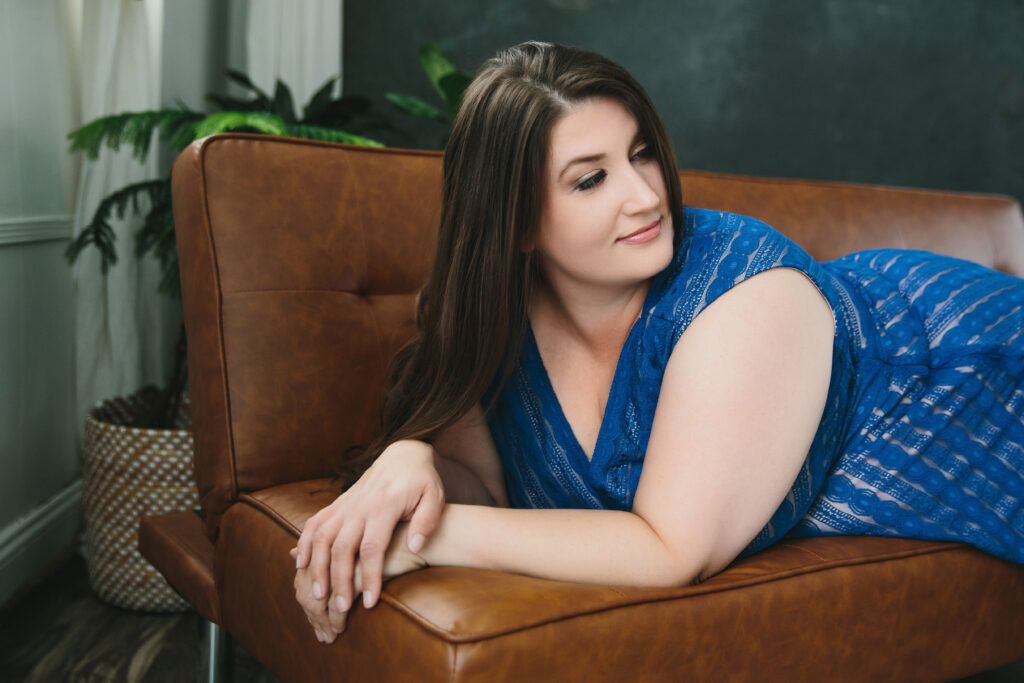 Woman in blue sundress leaning forward on a tan leather sofa. Photography by Lindsay Hite