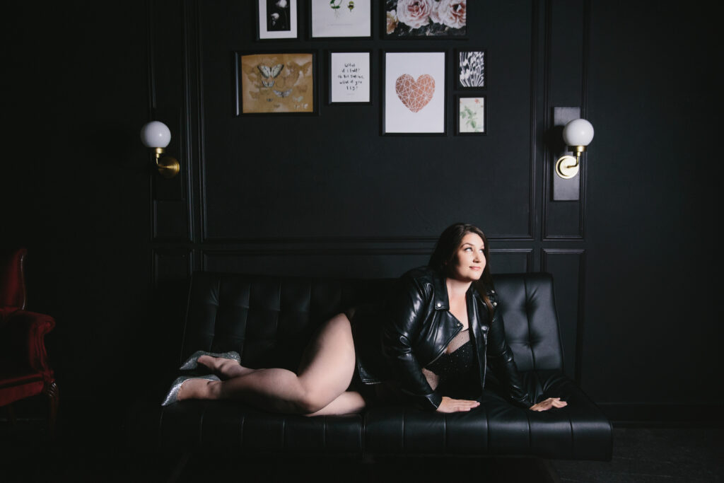 Woman in black body suit and black leather jacket wearing silver heels leans on a black sofa with a black backdrop displaying her confidence. Photography by Lindsay Hite