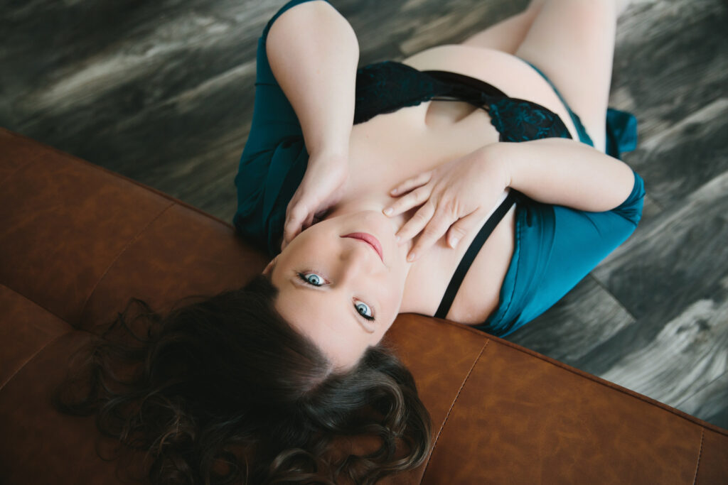 Woman in black bra and teal cardigan leaning back against a tan sofa looking at the camera. Photography by Lindsay Hite