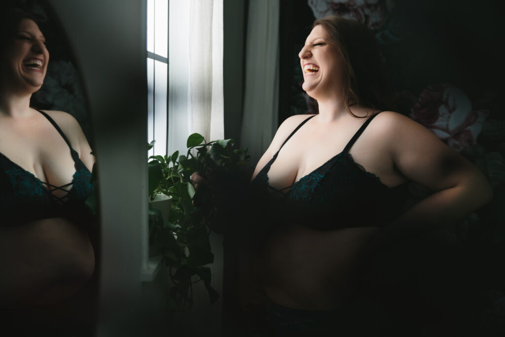 Woman laughing while looking out the window wearing a black bra. Photography by Lindsay Hite
