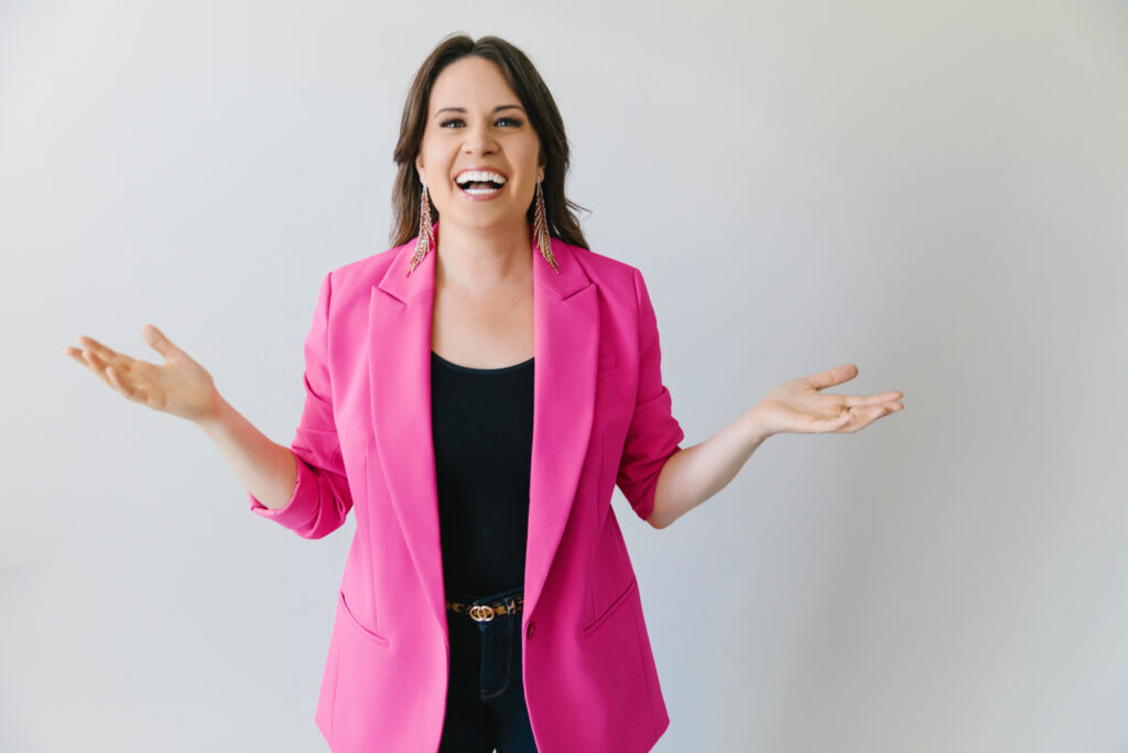 Woman wearing a black outfit with a bright pink blazer against a white background. Photography by Lindsay Hite