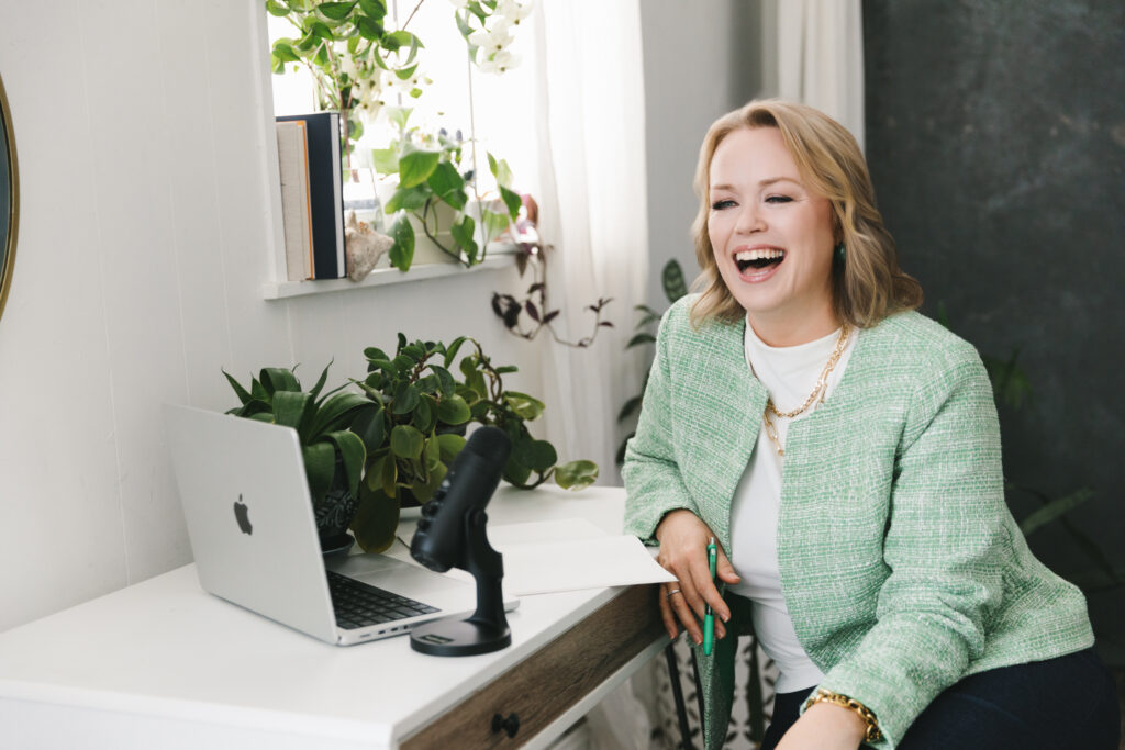Woman in a green blazer sitting at her desk looking at her computer with a microphone on the desk and greenery in the background. Photography by Lindsay Hite