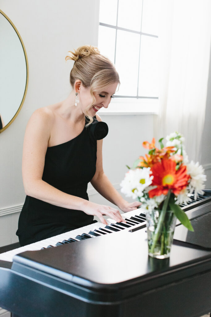 Woman wearing a one shoulder black jumpsuit playing the piano with flowers on the piano. Photography by Lindsay Hite