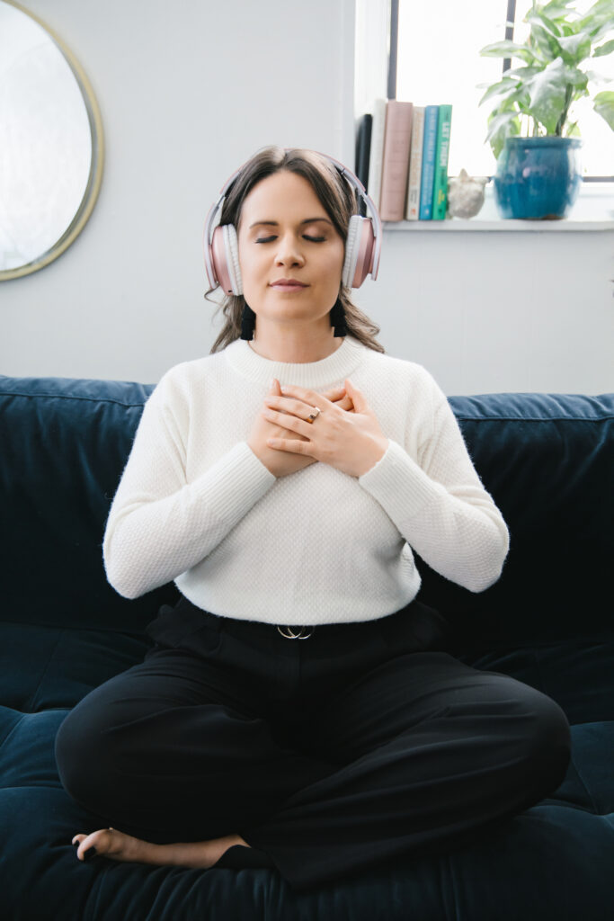Woman with a white sweater on, sitting cross-legged on a navy sofa wearing pink headphones with her eyes closed and hands over her heart. Photography by Lindsay Hit