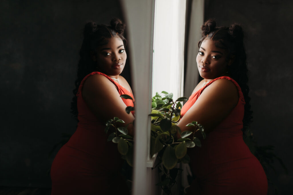 A woman in an orange dress leaning against a windowsill looking at the camera with her reflection showing in the mirror. Photography by Lindsay Hite