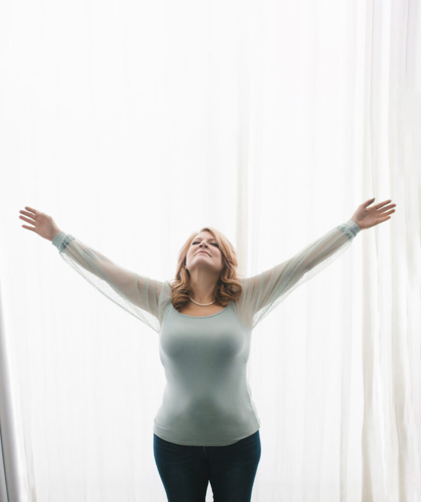 Woman in pale teal shirt looking up at the sky with her arms spread out wide in a star pose against a white curtain background. Photography by Lindsay Hite